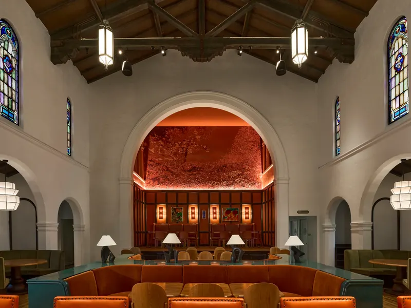 A vaulted dining hall with stained glass windows and long banquettes before a red arched alcove