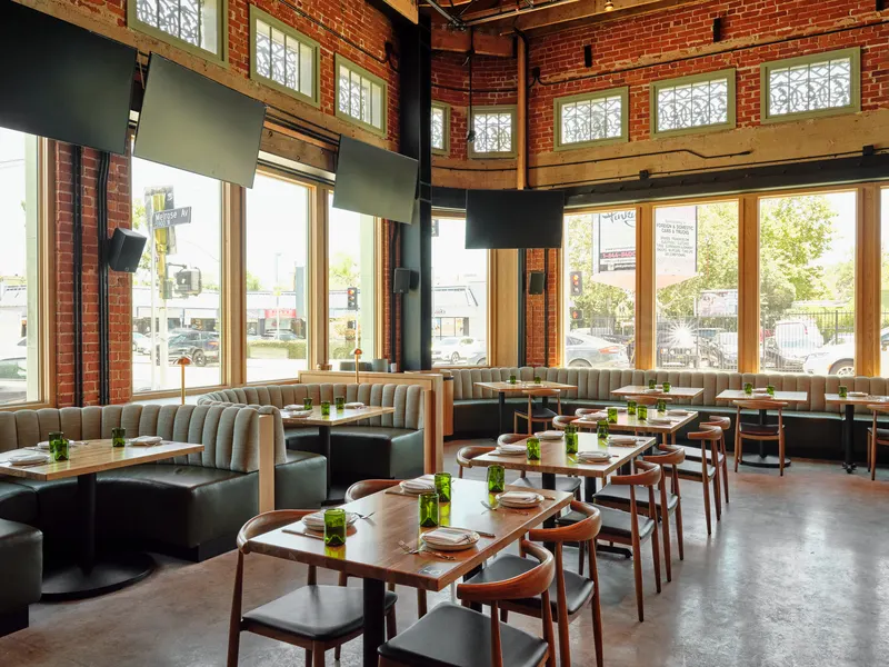 A dining room with green booths and timber tables, lit by tall windows in brick walls