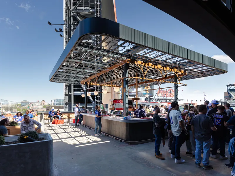 An open-air stadium deck with a bar under a steel canopy and a crowd of fans