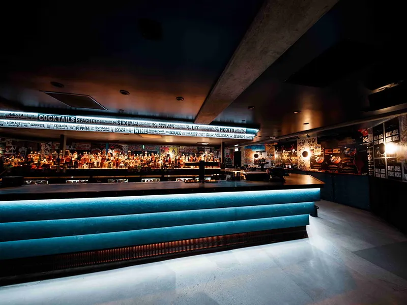 A long bar with a lit blue velvet front and a backlit drinks menu above the back bar