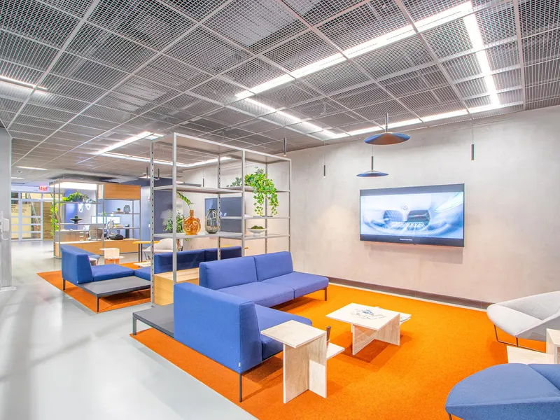 A showroom lounge with blue sofas on an orange rug under a metal ceiling grid