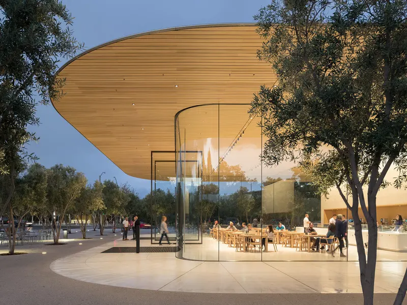 A glass pavilion under a timber soffit at dusk, with people at tables inside