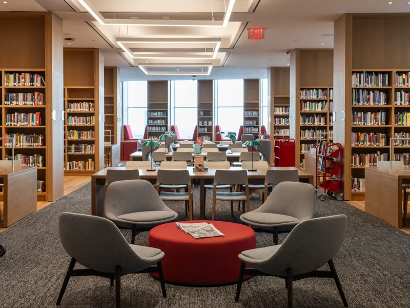 A library reading room with wood bookshelves, study tables and armchairs around a red ottoman