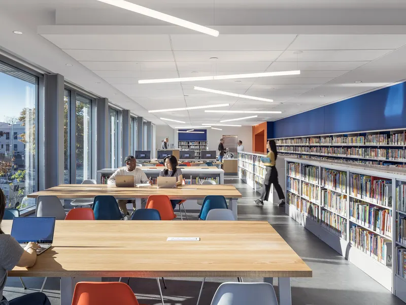 A library with long study tables, colored chairs and bookshelves along a window wall