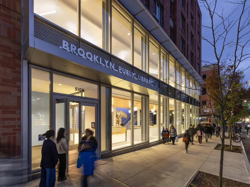 A glass library entrance at street level at dusk, with people walking past
