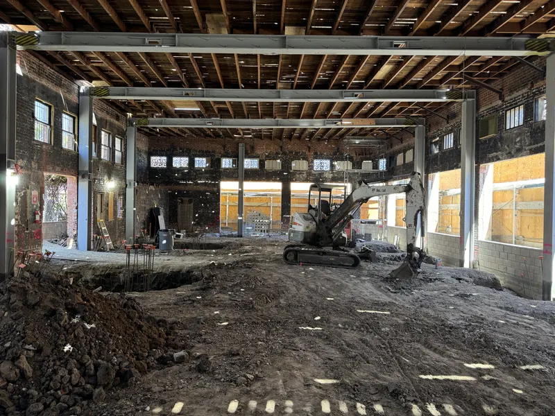 An excavator working on a dug-out floor inside a brick building under a timber roof