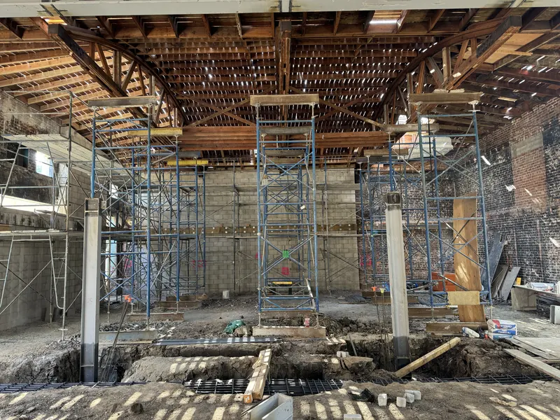 Shoring towers under a bow truss roof inside a brick building, with new footings dug into the floor