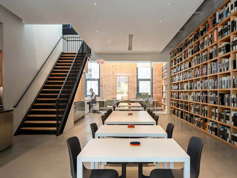 An office with long white tables, a black steel stair and a full-height bookshelf wall