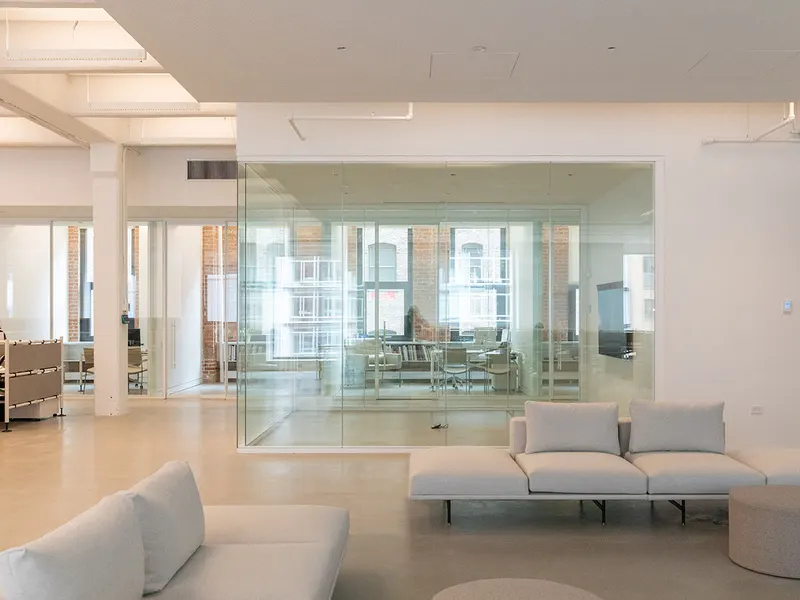 An office with a glass meeting room, white sofas and tall windows