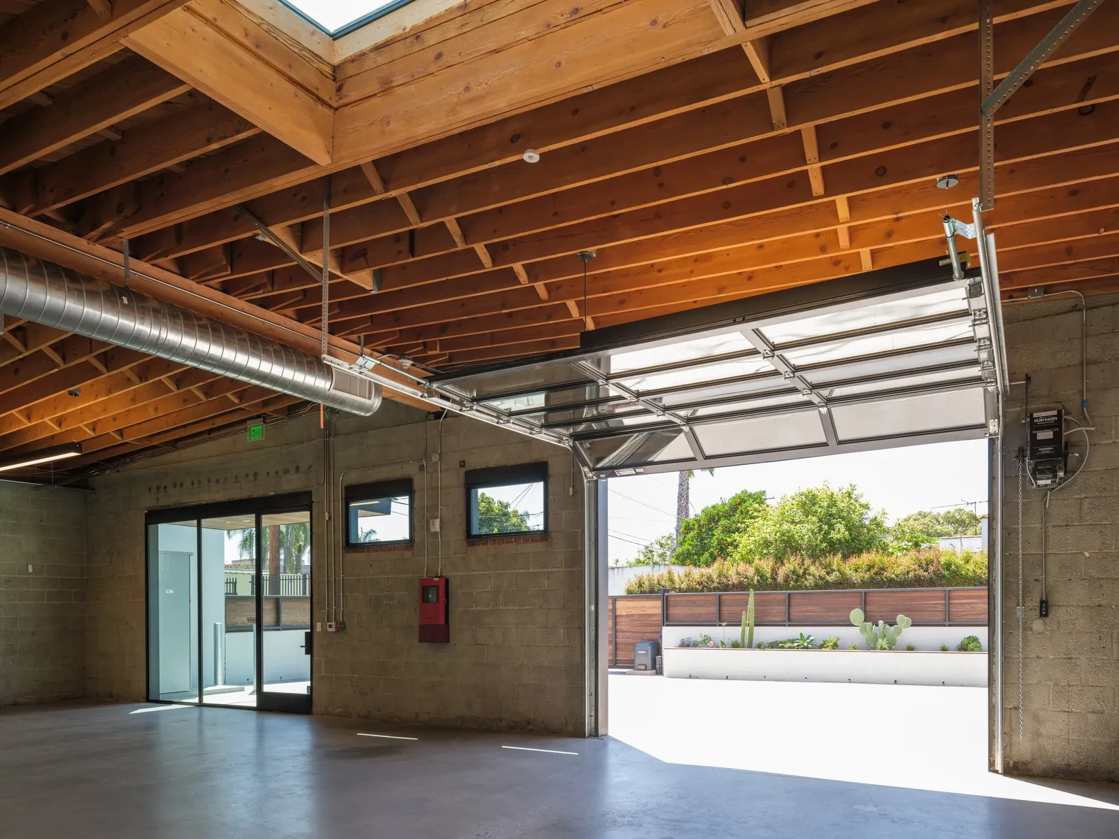 A block-walled office space under exposed timber joists, with a glass roll-up door open to a planted yard