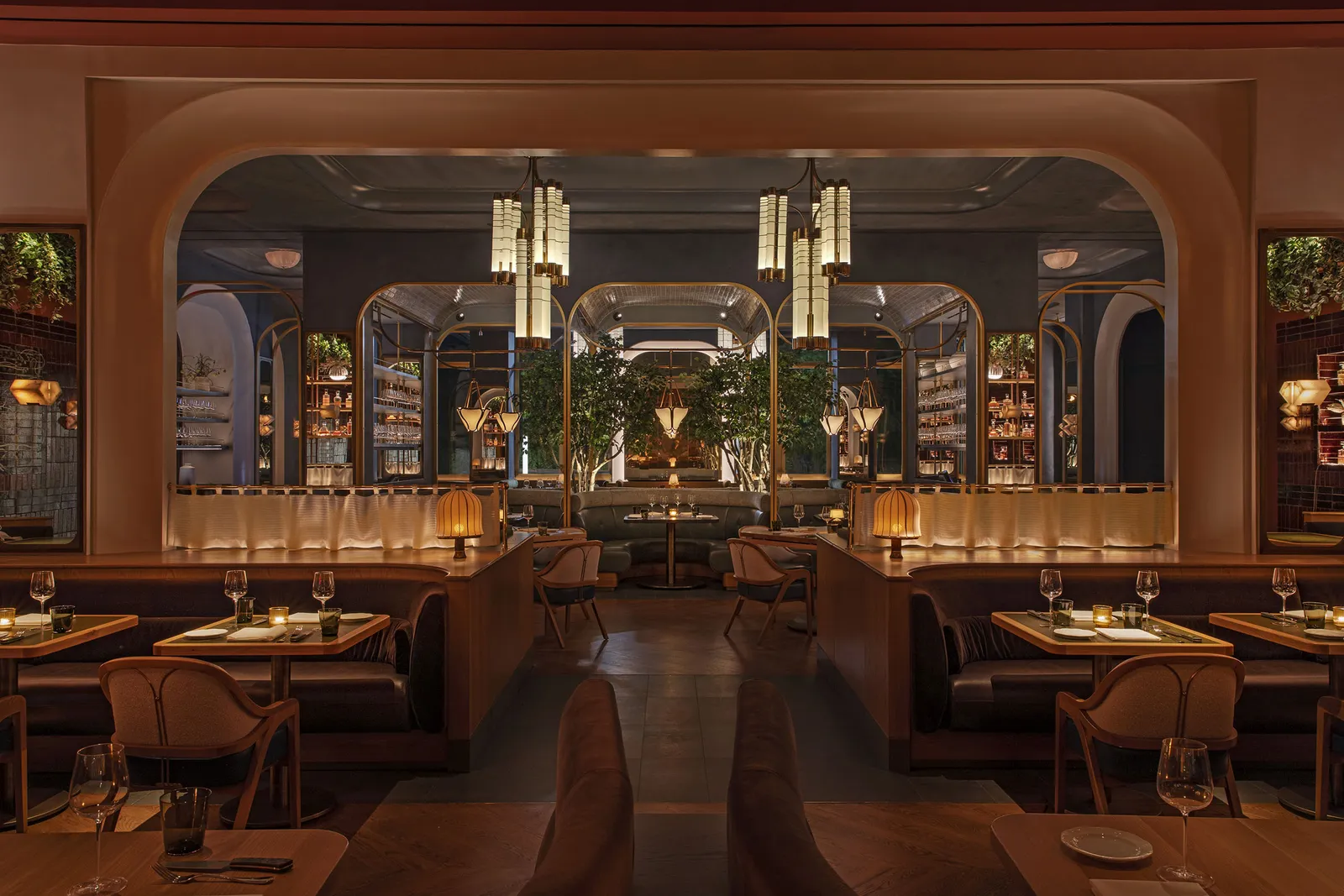 A dining room seen through a wide arch, with brass pendant lights, banquettes and trees beyond
