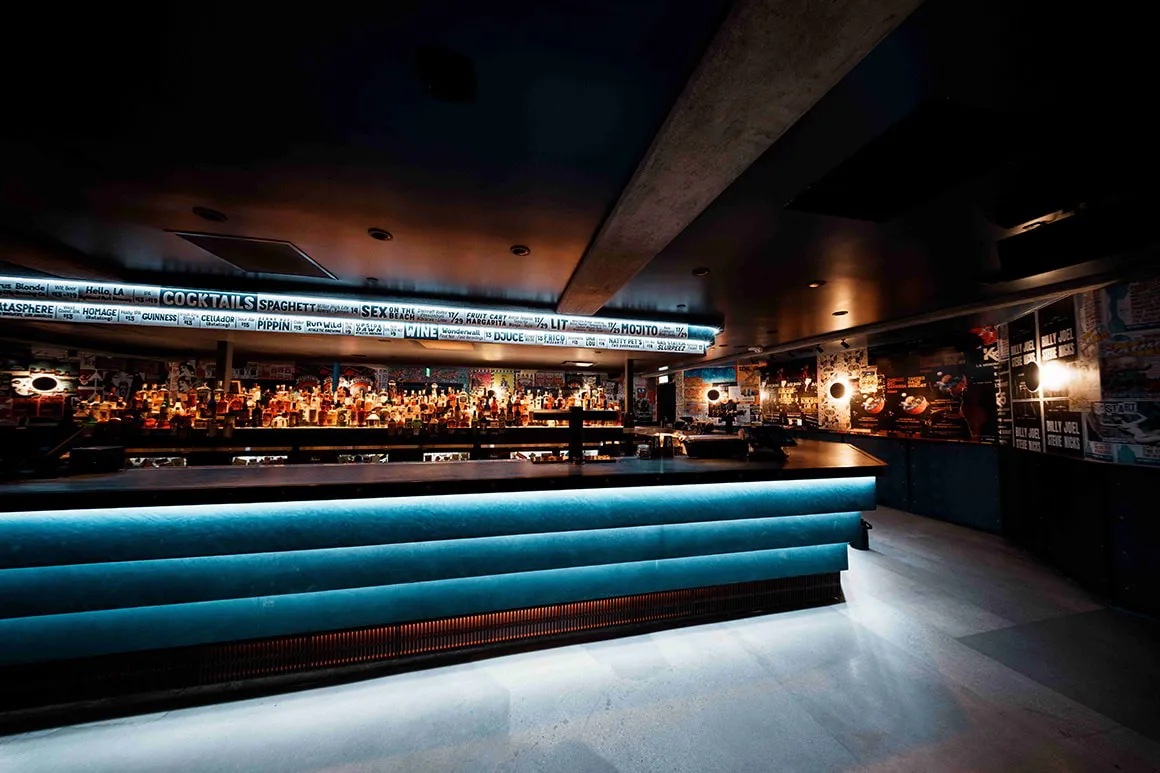A long bar with a lit blue velvet front and a backlit drinks menu above the back bar