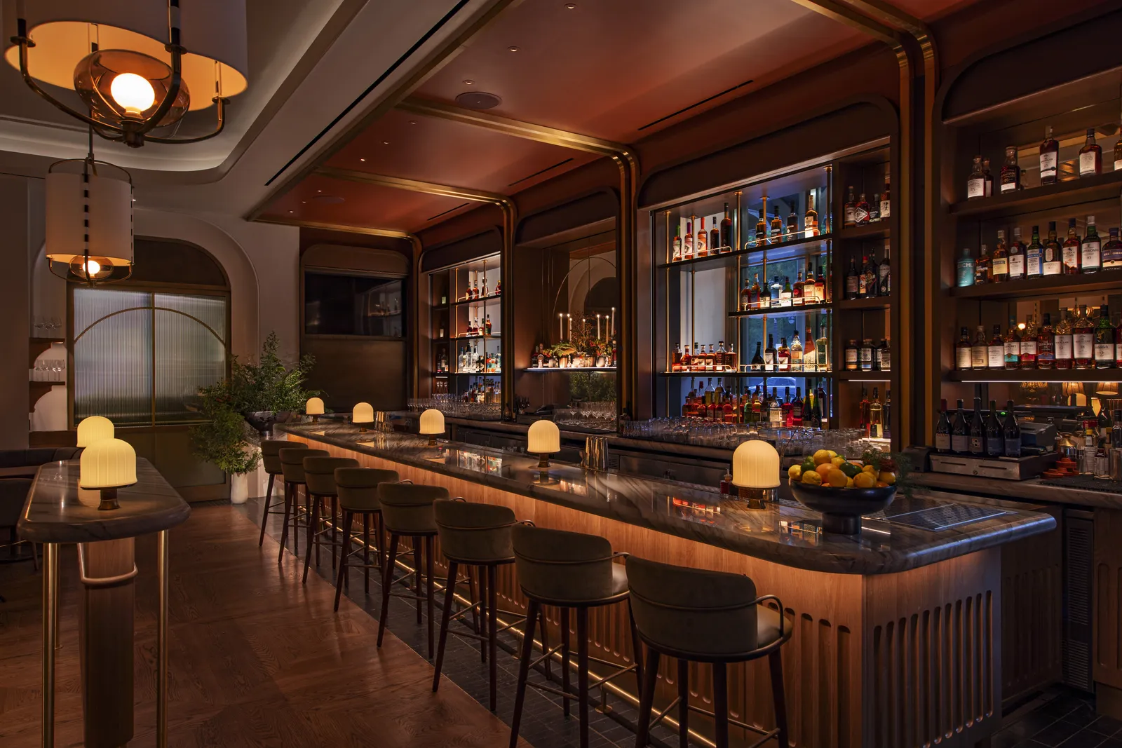 A restaurant bar with a marble counter, lit back shelving and table lamps along the bar